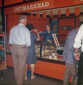 Några kunder står i kö till Ostmarknaden i Domus, Nya torget före 1976. En kvinnlig expedit står bakom ostdisken och låter en kvinna provsmaka.