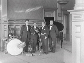 Gruppbild av orkester med kapellmästare Leo Pelez som poserar omgivna av sina instrument: trummor, cello, klarinetter, saxofon, fiol och flygel. (Se även bildnr GB2_110)