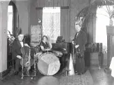 Lisa Lampas trio, bestående av två män och en kvinna, som sitter vid flygeln. Mannen till höger med fiol är Fritz Moreau och till vänster sitter en man med cello. Mellan herrarna står även trummor och blåsinstrument. Musikerna är fotograferade i matsalen på Varbergs Hotell. (Se även bildnr GB2_4013)