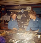 De kvinnliga expediterna Vally och Norma, iklädda blå rockar, står bakom fiskdisken, Domus vid Nya torget före 1976. I bakgrunden ses en våg och en apparat som visar NU BETJÄNAS 88 (könummer-system). På väggen sitter en stor svart/vit bild på ett fiskarsamhälle.