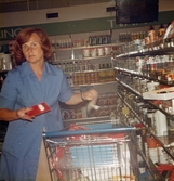 En kvinnlig expedit, iklädd blå rock, står bredvid en kundvagn och plockar ner/upp specerivaror i butiken, Domus vid Nya torget före 1976.