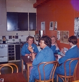 Ett kök/fikarum, Domus vid Nya torget senast 1976. Fyra kvinnor, iklädda blå rockar, sitter vid ett bord och fikar. På den röda/orangefärgade väggen bredvid kvinnorna sitter olika bilder uppsatta. I bakgrunden ses kökets brunmålade luckor och lådor.