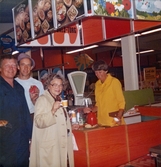 Till vänster ses en kvinnlig expedit, klädd i gul rock, som står bakom en röd-orangefärgad bröddisk där det bjuds på kaffe med tilltugg. På disken står en våg. Framför disken står tre kunder (två män och en kvinna) som ler mot fotografen. Den ene mannen håller upp en MER-förpackning (läskedryck) och kvinnan håller en pappmugg med kaffe i, Domus Nya Torget senast 1976.