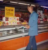 En ung man, klädd i blå rock och hållandes en pärm och penna i händerna, står vid en avlång orangefärgad kyl/frysdisk, Domus Nya Torget före 1976. I disken ligger förpackningar med spenat. Ovanpå disken står nyponjuice-förpackningar till försäljning. Strax bakom dem finns en gul skylt med texten: God idé - handla konsumentägt också i sommar.