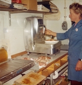 En kvinnlig expedit, klädd i blå rock, står i ett inplastat rum avsett för styckning av kött. På en arbetsbänk framför kvinnan står en våg samt styckade fläskbitar som ska vägas upp, Domus Nya torget före 1976.