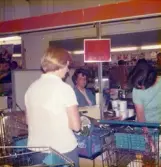 En kvinnlig expedit, klädd i blå rock, sitter i kassan och tar betalt, Domus Nya torget före 1976. Framför kassan står två kunder, en som packar varor och en som betalar.