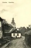 Stadsbebyggelse, Gamleby i Laholm med kyrktornet i bakgrunden. En man sitter på en sten intill den putsade husgaveln och vid knuten står ett litet barn.