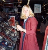 En kvinnlig expedit, klädd i röd rock, står i klädavdelning med papper och penna i handen, Domus på Frölundagatan efter 1976.