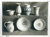 En skål med handtag, tre vaser, en kanna av modell 4011 samt två fat från Bo Fajans, tidigt 1900-tal. På bilden har dekoren på en vas delvis ritats i  med blå penna.