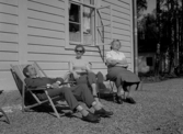 Två kvinnor och en man sitter intill en husvägg och vilar.