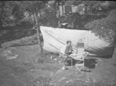 En man som sitter vid campingbordet och löser korsord med kaffepannan bredvid sig, samt lyssnar på transistorradion.
