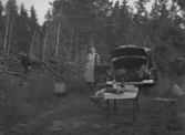 Ett campingbord står uppdukat med mat och dryck bakom en Volvo PV med bagageluckan öppen.