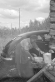 En kvinna står vid en husknut och förbereder diskning efter en god lunch, bakom henne en Volvo PV.