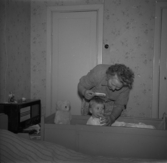 En mamma kammar sin lilla gosse där han sitter i sin barnsäng och hans lilla nallebjörn tittar nyfiket på.