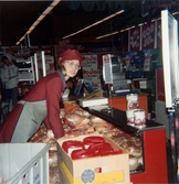 En kvinnlig expedit, klädd i röd rock och hätta samt grått förkläde, står i köttavdelningen och fyller på i kyldisken, Domus på Frölundagatan efter 1976.