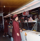 En kvinnlig expedit, klädd i röd rock och hätta, står innanför charkdisken och betjänar kunder. I bakgrunden skymtar en kvinna som förpackar en ost, Domus på Frölundagatan efter 1976.