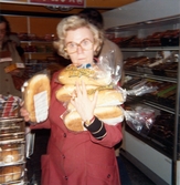 Berit Thörnqvist, klädd i röd rock, packar ner/upp bröd i brödavdelningen. Hon håller flertalet brödförpackningar i händerna, Domus på Frölundagatan 1976.