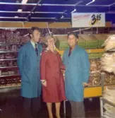 Tre personer står vid brödavdelningen, Domus Mölndal C 1976. Från vänster: Jan-Olof Standar (klädd i blå rock), Berit Thörnqvist (klädd i röd rock) och Kurt Ericsson (klädd i blå rock). Vid sidan av hänger en skylt i taket med texten 