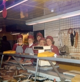 Fem kvinnor, klädda i röda rockar med hättor och grå förkläden, står innanför charkavdelningen, Domus Mölndal C 1976.