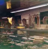 Fem kvinnor, klädda i röda rockar med hättor och grå förkläden, står innanför chark- och delikatessdisken, Domus Mölndal C 1976.