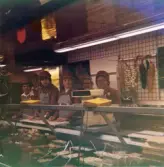 Fem kvinnor, klädda i röda rockar med hättor och grå förkläden, står innanför chark- och delikatessdisken, Domus Mölndal C 1976.