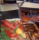Närbild i fiskdisken, Domus Mölndal C efter 1976. Till vänster ses en kokt hummer och en citron liggandes på grönsaker. Till höger ses en trälåda som innehåller rökt fisk. I bakgrunden ses inplastad lax, ål och tomater. Fiskdiskarna verkar vara separerade från varandra och den med hummern i är något högre placerad än den andra.