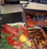 Närbild i fiskdisken, Domus Mölndal C efter 1976. Till vänster ses en kokt hummer och en citron liggandes på grönsaker. Till höger ses en trälåda som innehåller rökt fisk. I bakgrunden ses inplastad lax, ål och tomater. Fiskdiskarna verkar vara separerade från varandra och den med hummern i är något högre placerad än den andra.