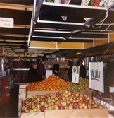 Stora fruktlådor fyllda med äpplen, apelsiner och päron för självplock står uppställda i frukt- och grönsaksavdelningen, Domus Mölndal C efter 1976. Bakom lådorna sitter en skylt med texten VÄG SJÄLV. I bakgrunden ses några kunder.