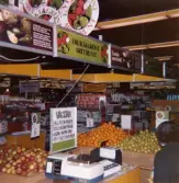 Stora fruktlådor fyllda med äpplen, apelsiner och päron för självplock står uppställda i frukt- och grönsaksavdelningen, Domus Mölndal C efter 1976. Vid lådorna står en våg samt en skylt med texten VÄG SJÄLV.