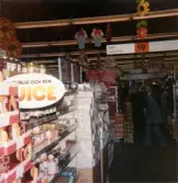 Till vänster ses en hylla med juice, sylt och konserver, Domus Mölndal C efter 1976. Längre bort till höger står en kvinnlig expedit, klädd i blå rock, och vinkar åt fotografen. I taket hänger en skylt med texten mjöl och sockerfritt (i gång 59).