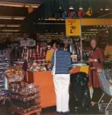 En kvinnlig demonstratris, klädd i gul rock, står vid en disk för provsmakning av bröd, Domus Mölndal C efter 1976. En kund och en expedit står framför disken. I bakgrunden ses stora, runda, uppstaplade aluminiumburkar med Cirkelkaffe.