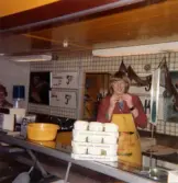 En kvinnlig expedit, klädd i röd rock och gult förkläde, står bakom fiskdisken hållandes en liten räka i händerna, Domus Mölndal C efter 1976. På disken ses förpackningar från Druvan, Eslöv, Smaksättare. På den vit-kaklade väggen i bakgrunden ses skyltar med olika sill-erbjudanden från Winner (Konsum-produkt).