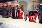 En man klädd i kavaj och slips, samt två kvinnliga expediter klädda i röda rockar, står innanför en disk, K-marknad Mölndals Centrum från 1986. I bakgrunden till vänster ses kosmetikaprodukter (Jane Hellen, Maxi, Invite och Pierre Robert) uppställda i hyllorna.