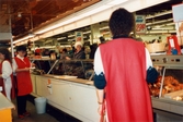 Tre kvinnliga expediter, klädda i röda rockar, står innanför köttdisken, K-marknad efter 1985/86. På utsidan disken står kunder.
