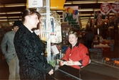 En kvinnlig expedit, klädd i röd rock, sitter i kassan och tar betalt, K-marknad efter 1985/86. En kvinnlig kund har lagt upp sina varor på bandet. Bakom henne skymtar en manlig kund.