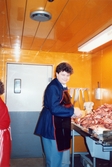 Styckmästaren står i ett orangefärgat inplastat rum avsett för styckning av kött, K-marknad i Mölndals centrum efter 1985/86. På diskbänken framför honom ligger uppstyckade köttdelar. I bakgrunden på väggen hänger sågar och knivar. Till vänster skymtas en kvinna.