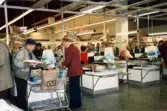 Tre kassor med dess personal som tar betalt vid rullbanden, K-marknad efter 1985/86. Butiken är full av kunder och en del lastar sina betalda varor i bruna papperskassar märkta med den blå Domus-symbolen (den liggande 8:an, oändlighetens symbol).