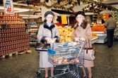 Två kvinnliga kunder, klädda i kappor, hattar och med handväskor, står med sina kundvagnar inne i butiken, K-marknad efter 1985/86. I bakgrunden ses upplagda frukter samt Cirkelkaffe till försäljning.