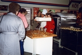En disk med nybakade wienerbröd står uppställd i butiken. Innanför disken står en kvinnlig expedit klädd i röd rock och en manlig bagare klädd i vitt. Bredvid kocken står en kvinna. I bakgrunden ses en stor ugn samt plåtar med förberedda bakverk som ska in i ugnen. Två kvinnliga kunder står framför disken och tittar på de nygräddade wienerbröden, K-marknad efter 1985/86.