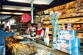 Två kvinnliga expediter (Berit Thörnqvist till höger) klädda i röda rockar står innanför bröddisken, Brödavdelningen på K-marknad, Mölndals C efter 1985/86. Hyllorna är fyllda av allehanda bakverk och en kund håller fram sin hand. På en stolpe sitter en röd kölapps-apparat.
