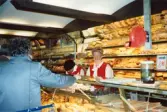 Två kvinnliga expediter klädda i röda rockar står innanför bröddisken, Brödavdelningen på K-marknad, Mölndals C efter 1985/86. Hyllorna är fyllda av allehanda bakverk och en kund håller fram sin hand. På en stolpe sitter en röd kölapps-apparat.