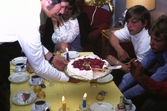 Tårtkalas hemma hos familjen Thörnqvist i Lindome, okänt årtal. Soffbordet är uppdukat med tända ljus och kaffeservice av finporslin. En man lutar sig fram över bordet och håller en tårta i händerna. Han bjuder två ungdomar att ta för sig.