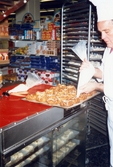 En manlig bagare klädd i vitt spritsar nybakade wienerbröd med sockerkristyr, K-marknad efter 1985/86. Till höger om honom står en ställning som innehåller flera plåtar med bakverk.