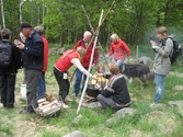 Besökare som deltar vid invigningen av Fagereds natur- och kulturstig i Lindome år 2008. Museitekniker Sven-Åke Svensson, klädd i röd jacka och blåjeans, står och bevakar elden där en stor järngryta hänger i en träställning ovanför. Till vänster ses en korg med ved och handgjorda dryckeskärl av trä. Personer i närheten äter ur små skålar. 
Fagered bjuder på kommunens bäst bevarade järnåldersmiljö. Här finns rester av en gammal hålväg, spår av äldre jordbruksmark och ett 40-tal gravar. 
Relaterade motiv: 2024_1300 - 1308.