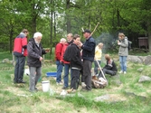 Besökare som deltar vid invigningen av Fagereds natur- och kulturstig i Lindome år 2008. Tredje person från vänster är museitekniker Sven-Åke Svensson, klädd i röd jacka och blåjeans, som står och bevakar elden. På marken ligger/står spänner. Personer i närheten äter ur små skålar. 
Fagered bjuder på kommunens bäst bevarade järnåldersmiljö. Här finns rester av en gammal hålväg, spår av äldre jordbruksmark och ett 40-tal gravar. 
Relaterade motiv: 2024_1300 - 1308.