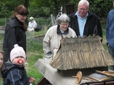 Besökare deltar vid invigningen av Fagereds natur- och kulturstig i Lindome år 2008. Ett litet barn sitter på knä framför en stenåldersby i mini-format (se 1988_0229). Byn består av ett hus, ställningar för att torka hud samt trädjur. Närmast barnet står museichef Mari-Louise Olsson (klädd i svart). I närheten står fler vuxna och beundrar den lilla byn. 
Fagered bjuder på kommunens bäst bevarade järnåldersmiljö. Här finns rester av en gammal hålväg, spår av äldre jordbruksmark och ett 40-tal gravar. 
Relaterade motiv: 2024_1300 - 1308.