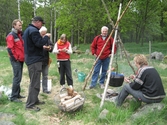 Besökare som deltar vid invigningen av Fagereds natur- och kulturstig i Lindome år 2008. Museitekniker Sven-Åke Svensson, klädd i röd jacka och blåjeans, står och bevakar elden där en stor järngryta hänger i en träställning ovanför. Till vänster ses en korg med ved och handgjorda dryckeskärl av trä. Personer i närheten äter ur små skålar. 
Fagered bjuder på kommunens bäst bevarade järnåldersmiljö. Här finns rester av en gammal hålväg, spår av äldre jordbruksmark och ett 40-tal gravar. 
Relaterade motiv: 2024_1300 - 1308.