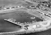 Flygbild över Glommens hamn med pir, fiskebåtar och mindre båtar, sjöbodar, magasin och omgivande enfamiljshus med trädgårdar. Fiskeläget utvecklades i samband med att hamnen anlades 1877-1880.