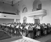 Kurs för lärarinnor i Rosenfredsskolans (Södra folkskolans) aula. Alla har var sin skolbänk med tvärslå att vila fötterna på. I bakgrunden står fyra män, möjligen kursledarna.