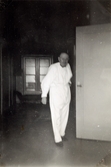 En vitklädd man (patient) håller i ett dörrhandtag i en korridor, Mölndals sjukhus 1939 - 1941. Bilder från fotoalbum, tagna från södra sidan av Länsmansgatan vid Mölndals lasarett/sjukhus 1939 - 1941 på bland annat givarens mor Ester (född Johansson) tillsammans med kollegan/väninnan Aina Englund. De arbetade på den manliga kirurgavdelningen som vårdbiträden/undersköterskor.
Relaterade motiv: 2024_1542 - 1617.